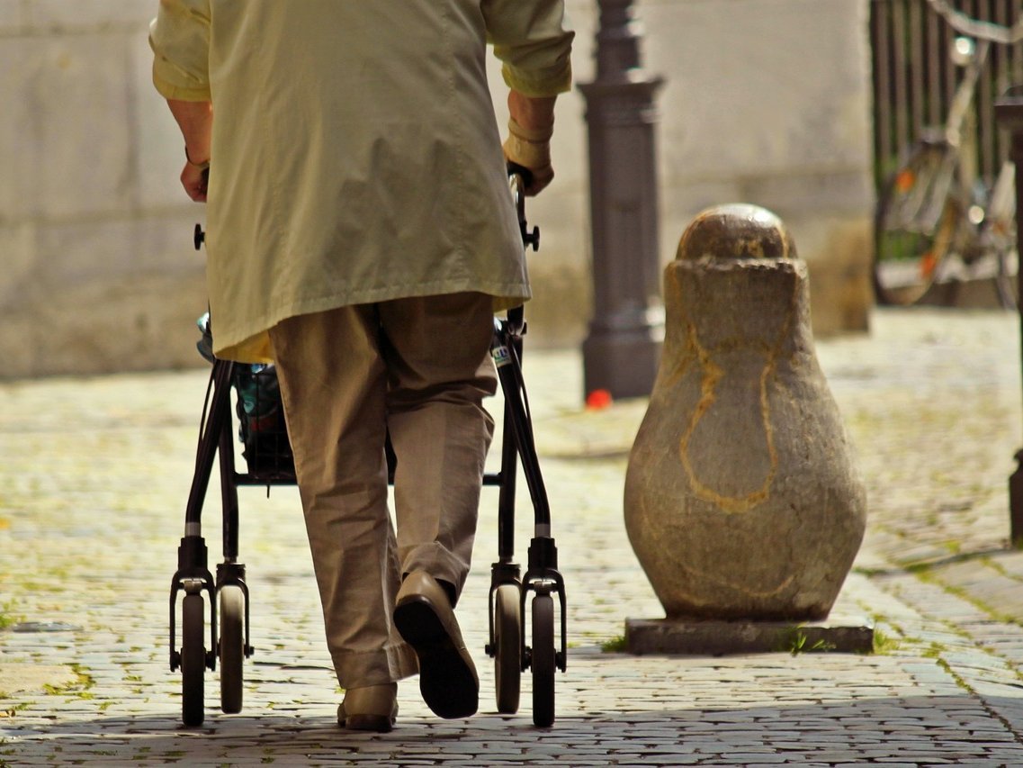 Eine Frau, die von hinten mit einem Rollator zu sehen ist, geht über eine Straße.